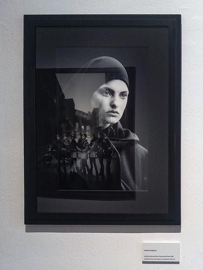 The picture shows a framed photo of a woman wearing a black headscarf. The glass reflects another photo on the opposite wall, which depicts 8 women in miniskirts.