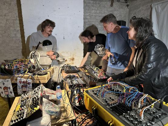 From right to left this photo includes Melissa Dyne, Dr.Spree, Robin Burke & Flo Flo. They're standing around 2 tables full of synthesizers with many cables visible, protruding from the modules that they are connected to. A table lamp is in front of Flo & illuminating the synth equipment.