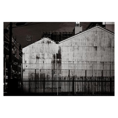This black-and-white photo features the corrugated metal side of a large industrial-style building in Nagoya. The twin gable roofs give the structure a mirrored, almost symmetrical appearance. Years of weathering have left long, dark streaks of rust and grime trailing down the metal siding, creating a pattern that looks both accidental and oddly deliberate. In the foreground, two layers of fencing—wire mesh and vertical bars—add a sense of separation or containment. The surrounding area includes glimpses of utility poles, wires, and nearby buildings, but the image is tightly composed to focus on the industrial wall, turning functional decay into graphic composition.