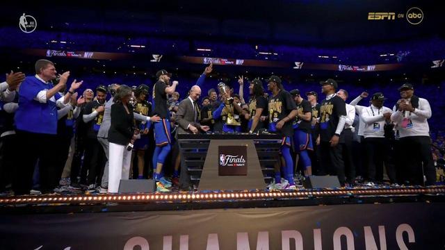 The Oklahoma City Thunder team stands on the championship stage as one player holds the Finals MVP trophy high. Teammates, coaches, and staff surround the stage wearing matching championship hats and shirts. The crowd in the background is fully engaged, filling the arena in Thunder blue.
