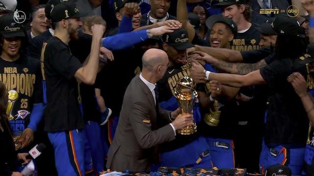A close-up shot of Finals MVP Shai Gilgeous-Alexander receiving the trophy from Adam Silver. His teammates enthusiastically gather around, reaching out to congratulate him. Shai’s expression is full of pride as he becomes the center of the celebration.