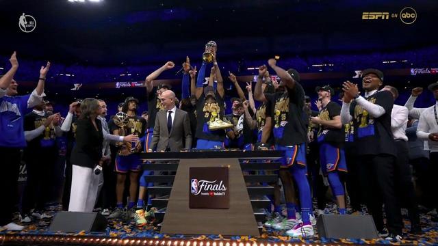 SGA, wearing the “NBA Champions 2025” t-shirt and team uniform, triumphantly raises the Larry O’Brien Championship Trophy above his head at center stage. Confetti litters the stage floor as teammates surround him in celebration, with commissioner Adam Silver and media members to the side.