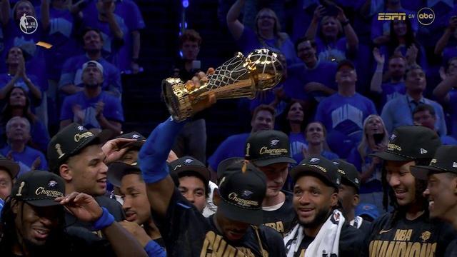 Shai Gilgeous-Alexander raises the NBA Finals MVP trophy high above his head, surrounded by smiling teammates. Fans in the background, clad in Thunder blue, cheer and capture the moment on their phones. Shai’s confidence and joy are evident as he shows off the golden prize.