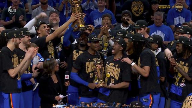 A jubilant Oklahoma City Thunder team celebrates their NBA Championship victory on the court. Players in black “NBA Champions 2025” t-shirts and black caps lift three trophies high: the golden Larry O’Brien Championship Trophy, the Bill Russell NBA Finals MVP Trophy, and a third golden commemorative basketball trophy. The players are smiling, shouting, and pointing in all directions, surrounded by cheering teammates. One player holds both the MVP and commemorative trophies, beaming with pride, while another holds the Larry O’Brien Trophy above his head. Confetti covers the stage, and fans in blue shirts applaud in the background.