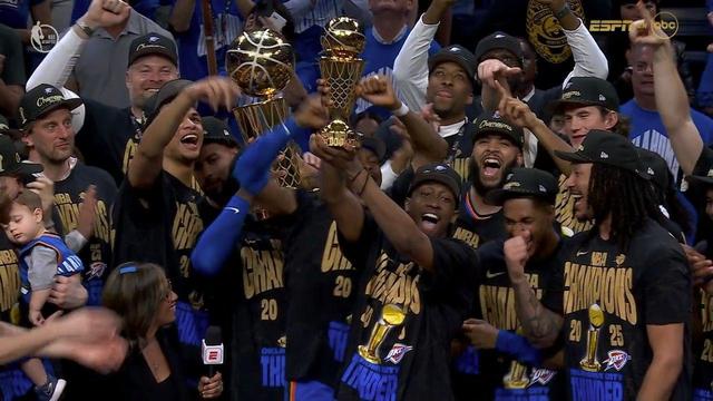 An Oklahoma City Thunder player lifts the Larry O’Brien Trophy high above his head as the team celebrates their 2025 NBA Championship win. Another player, smiling widely, holds the Finals MVP Trophy at chest level. Teammates cheer, clap, and point excitedly, many still wearing their black and gold “NBA Champions 2025” shirts and black caps. An ESPN reporter stands in front of the group with a microphone. One player cradles a baby in Thunder gear. The mood is euphoric, with everyone on stage caught in the moment of triumph.