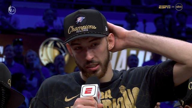 Chet Holmgren from the Oklahoma City Thunder is being interviewed courtside after winning the NBA Finals. He wears a black “Champions 2024/2025” hat with the OKC logo and a matching NBA Finals Champions shirt with gold graphics. He looks reflective and slightly emotional, with his right hand placed behind his head. He holds a microphone with the ESPN logo in his left hand. The arena behind him is lit with purple and gold hues, and fans can be seen smiling and clapping. The broadcast overlay shows the ESPN and ABC logos in the top-right corner.