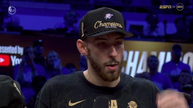 Chet Holmgren from Oklahoma City Thunder player continues the post-game interview. He now has both hands down and is speaking into the ESPN microphone with a focused, calm expression. He wears the same black “Champions 2024/2025” hat and NBA Finals Champions shirt. The background features a crowd of fans and bold gold “CHAMPIONS” signage celebrating the team’s title win. The lighting remains dramatic, and the ESPN on ABC overlay is visible in the top-right corner.