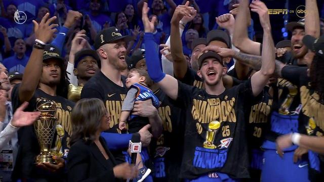 The Oklahoma City Thunder celebrate their 2024–2025 NBA Championship on the court. A joyful crowd of players, wearing black and gold “NBA Champions 2025” shirts and hats, raise their arms and cheer. One player cradles a baby dressed in a miniature Thunder jersey, adding a heartwarming moment to the celebration. Another player holds the Larry O’Brien Championship Trophy. An ESPN reporter is present with a microphone, capturing the postgame excitement. Fans and teammates in the background smile and clap amid the festive atmosphere, all under the arena’s bright lights.