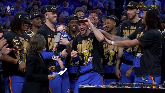 The Thunder’s championship celebration continues as players gather closely, laughing and smiling for the cameras. One player holds a baby in a Thunder jersey, who appears to be napping during the excitement. Another player tips his championship hat and beams with joy. The Larry O’Brien trophy is visible again, held proudly by a teammate. An ESPN reporter stands nearby, preparing for interviews. Confetti and decorations cover the foreground table. The atmosphere is filled with camaraderie and pure joy after their Finals victory.