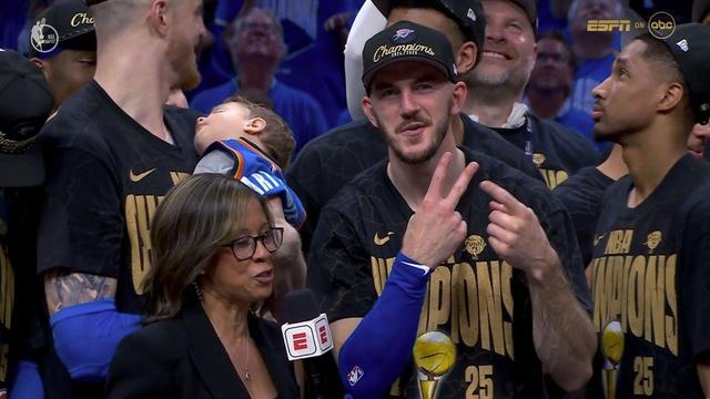 Oklahoma City Thunder player Alex Caruso is being interviewed courtside after winning the 2024–2025 NBA Championship. Wearing a black “Champions 2024/2025” hat and NBA Champions shirt, Caruso flashes two fingers in a victory gesture while smiling at the camera. A reporter with an ESPN mic stands in front of him, and behind them, a teammate cradles a sleeping baby in a tiny Thunder jersey. Other players in matching gear stand around them, beaming and enjoying the celebration.