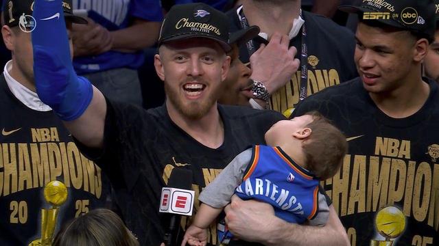 Isaiah Hartenstein, Oklahoma City Thunder player celebrates the team’s 2024–2025 NBA Championship on court, smiling widely and raising one arm in the air. He wears a black “Champions 2024/2025” hat and matching black and gold “NBA Champions 2025” shirt. In his other arm, he cradles his son Wyatt wearing a custom Thunder jersey that reads “HARTENSTEIN.”He appears to be peacefully asleep amid the excitement. Other Thunder players in championship gear surround him, smiling and watching the moment. An ESPN microphone is visible near the player’s chest as the postgame interviews continue.