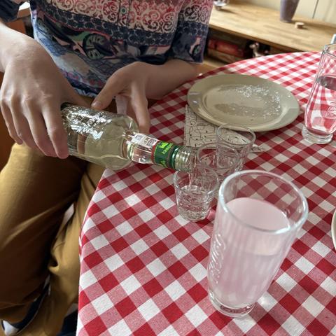 Can see a pair of hands pouring vodka into three little shot glasses on a table covered with red and white cloth. There is a taller glass filled with water and an empty plate on the side. The person pouring has mustard colour pants and a shirt with red and blue intricate patterns.