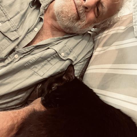 a black cat sleeps against the arm of a smiling man on a couch