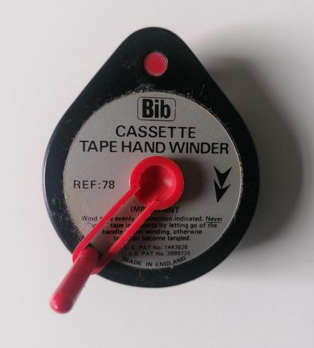 Bib cassette tape hand winder. A black plastic tear shaped object with a red plastic crank handle. A silver circular label with black lettering has the branding and title plus arrow heads to indicate direction the handle should be turned