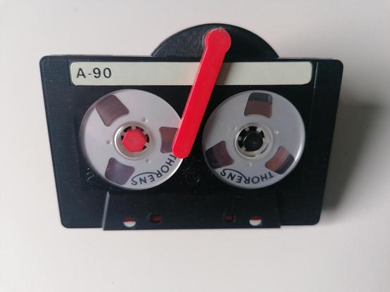 A black shell cassette tape with 2 circular transparent windows reveling a mock reel to reel metallic-like reels inside, branded Thorens on the metal reels. A red arm clamps the cassette to a manual tape rewind mechanism