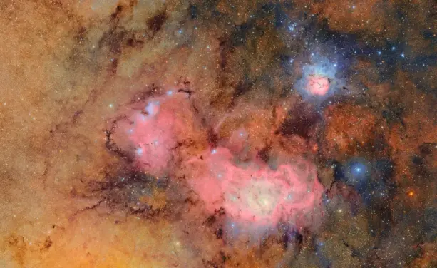 A cosmic tapestry of glowing tan and pink gas clouds with dark dust lanes. In the upper right, the Trifid Nebula resembles a small flower in space. To the lower left, the larger Lagoon Nebula stretches wide like a churning sea of magenta gas, with bright blue, knotted clumps sprinkled throughout. 