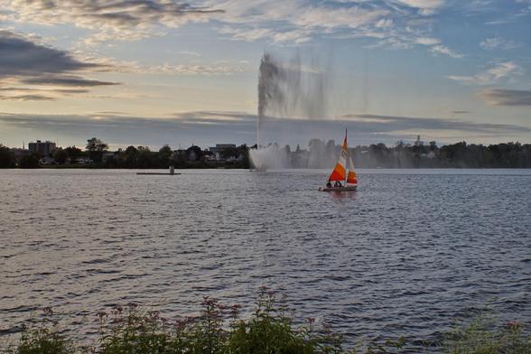 A small sailboat with orange and white sails glides past a large fountain in a lake. The late evening sky glows from the setting sun highlighting the clouds. Trees can be seen on the distant shoreline and some plants line the nearby shore.