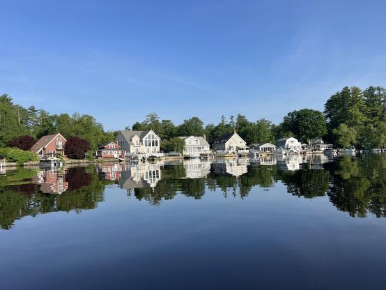 Five houses lined up on a shore with trees behind them. Their reflection is shown on a smith as glass lake. 