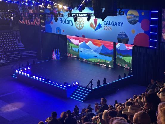 A colorful stage setup for the Rotary event is displayed, featuring a backdrop with nature-themed graphics and the text "Calgary 2025." Flags are positioned on stage, and an audience is visible in the foreground, with some attendees wearing cowboy hats.