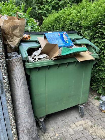 Grüne Bio Tonne voll mit Pappe und Papier. Daneben ein alter zusammengerollter Teppich.