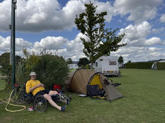 Een man in een geel shirt zit op een ligfietstrike voor een pol siergras en ernaast staat een groene tunneltwnt met de deur open op het groene gras. Erachter staat een boom en een witte caravan. De lucht is blauw met witte wolken