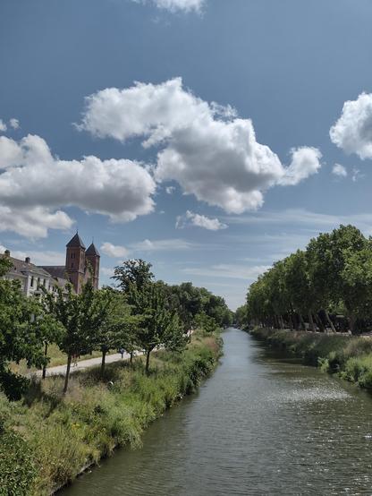 Foto genomen vanaf een brug. Een kanaal met aan de rechterkant van het kanaal hoge bomen en aan de linkerkant kleine bomen, een wandelpad en daarachter een kerk. Verder weg staan links en rechts hoge bomen.