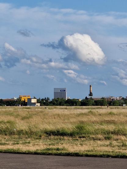 Blick über das Tempelhofer Feld nach Süden. Ein Fabrikgebäude weiter südlich hat eine lichtgraue Fassade in der obersten Etage, welches fast durchsichtig erscheint bei Sonnenlicht. Und so den Baumwipfel und Schornstein dahinter schweben lässt.
