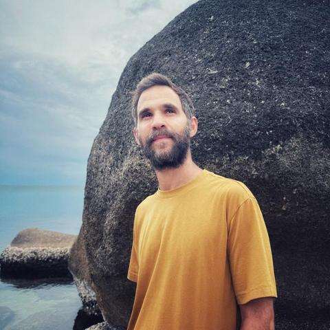 Kutiman wears a calm expression and a yellow t-shirt,  standing by a large rock near the sea, under an overcast sky.
