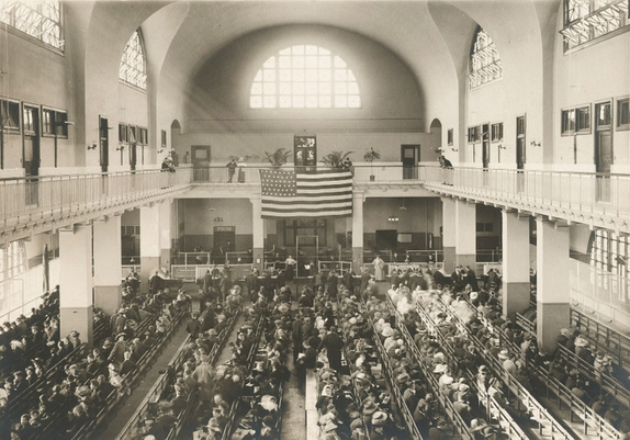Photo of the interior of the main hall at the Ellis Island processing center, circa 1900, showing immigrants seated on rows of benches, with a large American flag on the center far wall. 