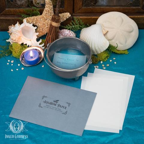 Three sheets of "AquaVow Paper" product are arranged on the right in front of a small silver cauldron, which contains blue water and a slip of paper with the words "money flows to me effortlessly" on it. An envelope stamped with "AquaVow Paper / Make a Sacred Vow, Let the Water Set it Free" sits to the left. Presented by Inked Goddess Creations.