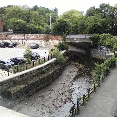 Photograph of the tidal stretch of Ouseburn River at south side of Ouseburn Bridge in Newcastle upon Tyne. To the left (south-west) of the bridge is Cut Bank, and to the right (north-east) is Byker Bank. In the foreground is the pedestrian Riverside Walk and the photo also spans some surrounding built-up areas and trees, with sky in the background. Still image captured from webcam at 20:28 hrs on Monday 23 June 2025.