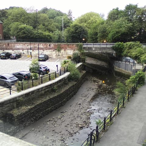 Photograph of the tidal stretch of Ouseburn River at south side of Ouseburn Bridge in Newcastle upon Tyne. To the left (south-west) of the bridge is Cut Bank, and to the right (north-east) is Byker Bank. In the foreground is the pedestrian Riverside Walk and the photo also spans some surrounding built-up areas and trees, with sky in the background. Still image captured from webcam at 20:52 hrs on Monday 23 June 2025.