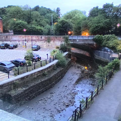 Photograph of the tidal stretch of Ouseburn River at south side of Ouseburn Bridge in Newcastle upon Tyne. To the left (south-west) of the bridge is Cut Bank, and to the right (north-east) is Byker Bank. In the foreground is the pedestrian Riverside Walk and the photo also spans some surrounding built-up areas and trees, with sky in the background. Still image captured from webcam at 21:49 hrs on Monday 23 June 2025.