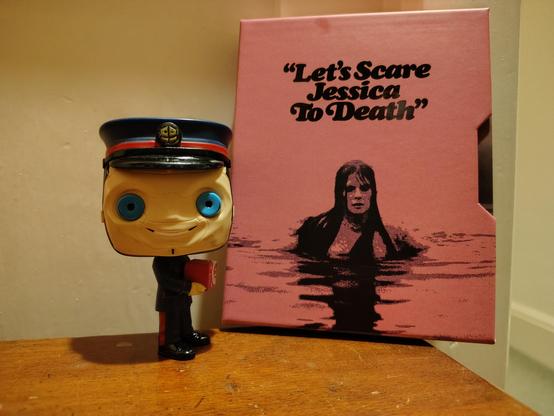 The Kerblam Man figure from Doctor Who standing next to a new 4K disc edition of the 1971 independent horror movie Let's Scare Jessica To Death.
