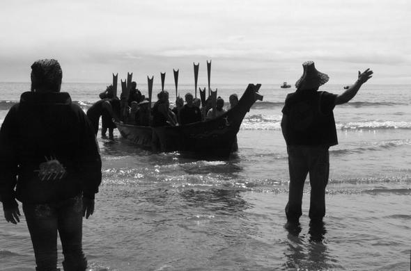 A large red cedar dugout canoe with many paddlers, their paddles held with blades raised, rests just offshore as a tribal elder standing on the shallows and wearing a woven cedar bark hat raises his hand while talking story. It's a black and white photo, and another tribal members stand at left. The bow of the canoe is topped with a carving that appears to be a wolf head. The ocean is calm and the skies overcast and gray.