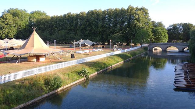 The College Backs in Cambridge looking out from Garret Lane Bridge towards Trinity Bridge. On the right punts are lined up by the side of the river. On the left the sun is shining on awnings, tents and various constructions that make up May Ball paraphernalia in the Trinity paddocks, all apparently deserted.