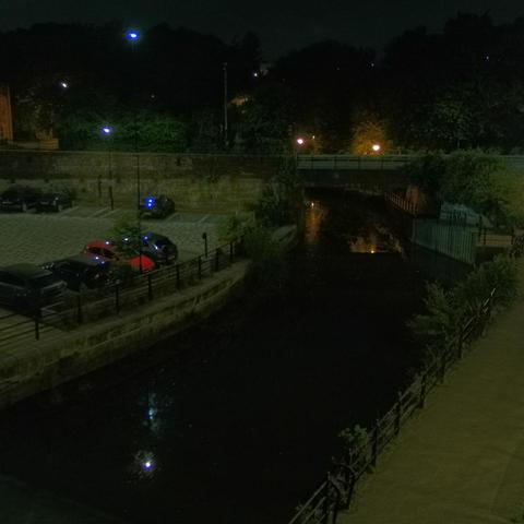 Photograph of the tidal stretch of Ouseburn River at south side of Ouseburn Bridge in Newcastle upon Tyne. To the left (south-west) of the bridge is Cut Bank, and to the right (north-east) is Byker Bank. In the foreground is the pedestrian Riverside Walk and the photo also spans some surrounding built-up areas and trees, with sky in the background. Still image captured from webcam at 02:32 hrs on Tuesday 24 June 2025.