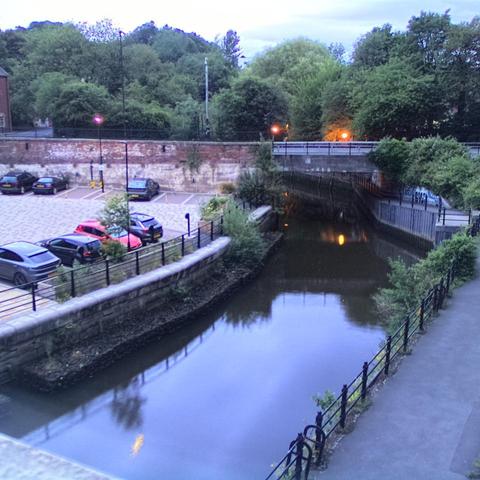 Photograph of the tidal stretch of Ouseburn River at south side of Ouseburn Bridge in Newcastle upon Tyne. To the left (south-west) of the bridge is Cut Bank, and to the right (north-east) is Byker Bank. In the foreground is the pedestrian Riverside Walk and the photo also spans some surrounding built-up areas and trees, with sky in the background. Still image captured from webcam at 04:24 hrs on Tuesday 24 June 2025.