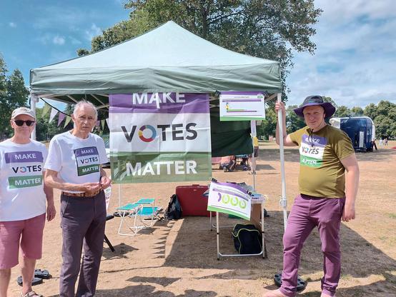 3 people in front of a stall with a large "Makes Votes Matter" poster