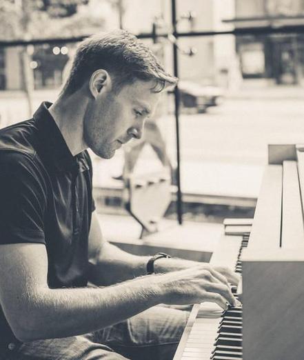 A black and white photo of Dale Nicols, the Vancouver pianist and composer, playing piano in a quiet, sunlit room. The atmosphere is calm and focused, with a cityscape visible through large windows. 
