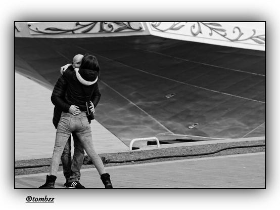 Black and white photo of a couple in a long kiss, standing on a waterfront. The woman is wearing a dark jacket, light fitted trousers, and dark shoes. The man is dressed in a dark jacket, dark trousers, and shoes. Their bodies are entwined in a close embrace, capturing an intimate moment. In the background, there’s a glimpse of the sea and the stern of a moored sailing ship.