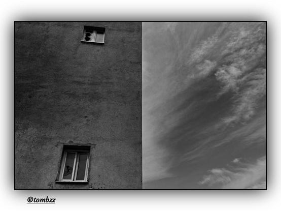 A black and white photograph divided vertically into two parts: on the left side, a fragment of an old building with two windows, their glass shattered; on the right side, clouds stretching across the sky. The contrast between the ruin and the open sky gives the image a symbolic dimension.