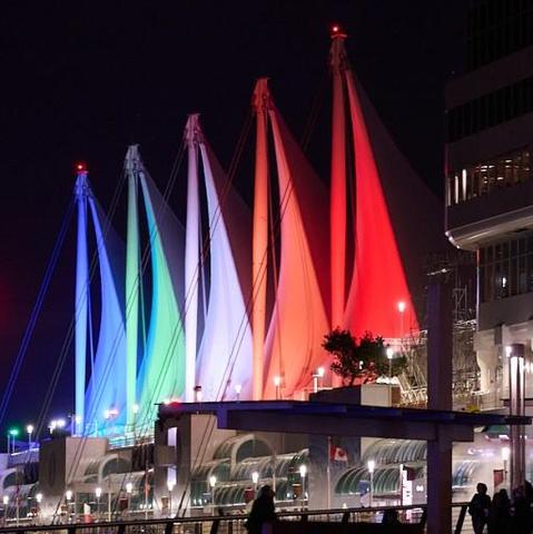 Sails looking like flags in different colours lit up from below at night in front buildings with people in silhouette.
