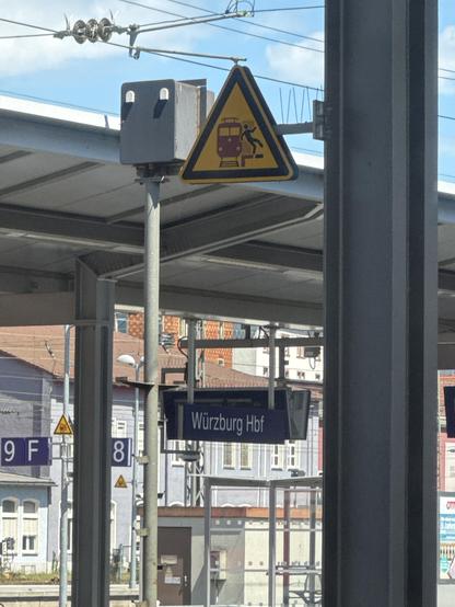 Das Bild zeigt eine Schilderung am Würzburg Hauptbahnhof (Hbf). Im Vordergrund ist ein gelbes Warnschild mit einem schwarzen Rand zu sehen, das eine Person, die auf die Bahn zustürzt, darstellt. Dieses Schild dient als Warnung für Passagiere, um auf die Gefahr von Stürzen auf der Bahn zu achten. Direkt unter dem Warnschild hängt ein blaues Schild mit der Aufschrift "Würzburg Hbf", das den Bahnhof identifiziert. Im Hintergrund sind Teile des Bahnhofsgebäudes sowie einige Gebäude im Stadtbild zu …