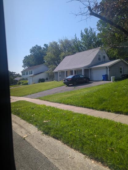 A gray cape cod style house with an attached garage. I the driveway is a black Toyota Corolla from the early 2000s. A sizable green lawn is in the foreground.