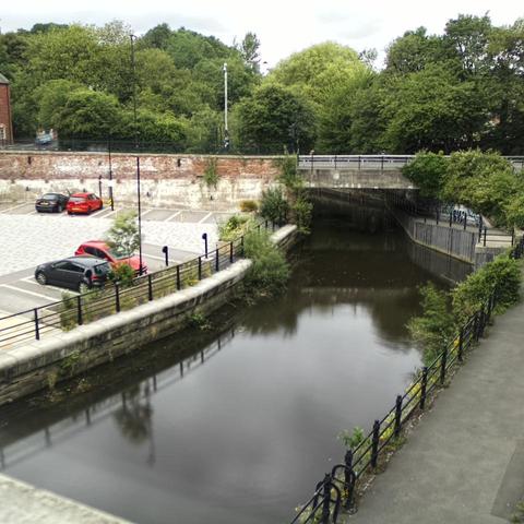 Photograph of the tidal stretch of Ouseburn River at south side of Ouseburn Bridge in Newcastle upon Tyne. To the left (south-west) of the bridge is Cut Bank, and to the right (north-east) is Byker Bank. In the foreground is the pedestrian Riverside Walk and the photo also spans some surrounding built-up areas and trees, with sky in the background. Still image captured from webcam at 15:17 hrs on Tuesday 24 June 2025.
