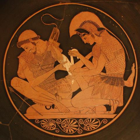 Red-figure vase painting of Achilles, depicted as a beardless young man, bandaging Patroclus, who is depicted as a mature bearded man. Both are still in full armour, wearing helmet, cuirass (to protect the torso) and pteryges, the "leather skirt" so iconic in Greek armour. Patroclus is sitting with spread legs, exposing his flaccid penis, giving the scene an erotic element.