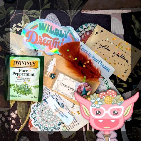Photo of the contents of a Goblin Dross package, all ready to be put in an envelope and mailed out. It includes a packet of peppermint tea, a shiny sticker that reads "wildly dreaming", a brown feather, a small white plastic figure of a woman in a dress, a round mandala-style sticker, several partially obscured art inspiration cards, a paper slip fortune from a fortune cookie, and a one of a kind one-inch button with a star and mushroom design - attached to a piece of paper that reads "navigate." There is also a brown paper envelope with "golden glitters" handwritten on its face, and a few gold heart confetti pieces sprinkled over everything. At the bottom right of the image is an illustration of a smiling pink goblin with white glasses and a purple hoodie, wearing flowers and mushrooms and a snail shell on her head.