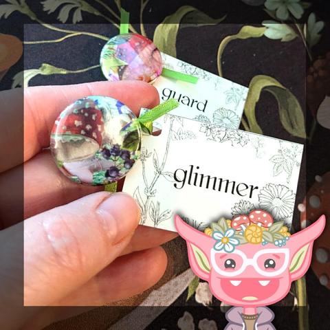 Close-up of two of the one-of-a-kind, handmade one-inch button fortunes. One in the background that reads "guard," and one being held up closer by a hand that reads "glimmer." At the bottom right of the image is an illustration of a smiling pink goblin with white glasses and a purple hoodie, wearing flowers and mushrooms and a snail shell on her head.