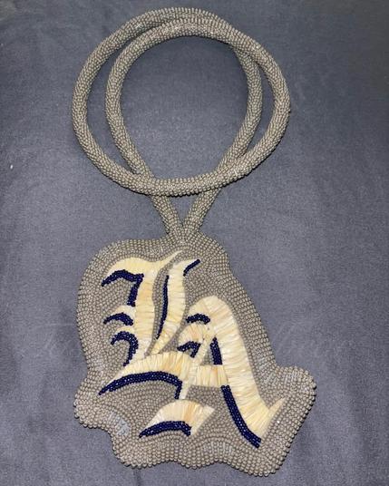 a beaded gray medallion made by either kiamichi or her daughter with the letters LA in blue and white in the center in old english font made from porcupine quills. resting on a dark gray background